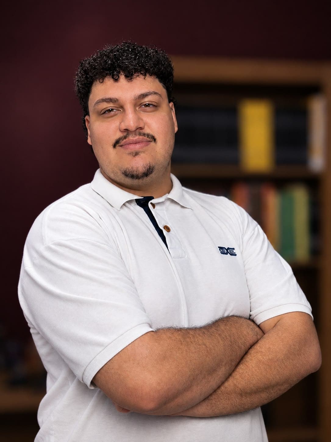 Retrato de Julio Paes Jr., de braços cruzados, com estante de livros ao fundo