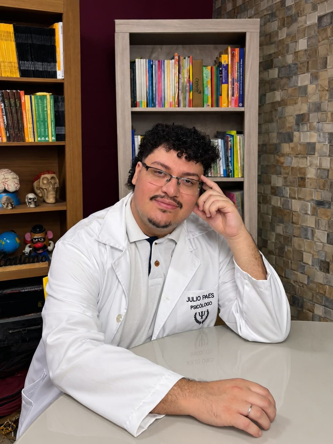 Julio Paes Jr. sorrindo, com a mão apoiada no rosto, sentado à mesa do consultório
